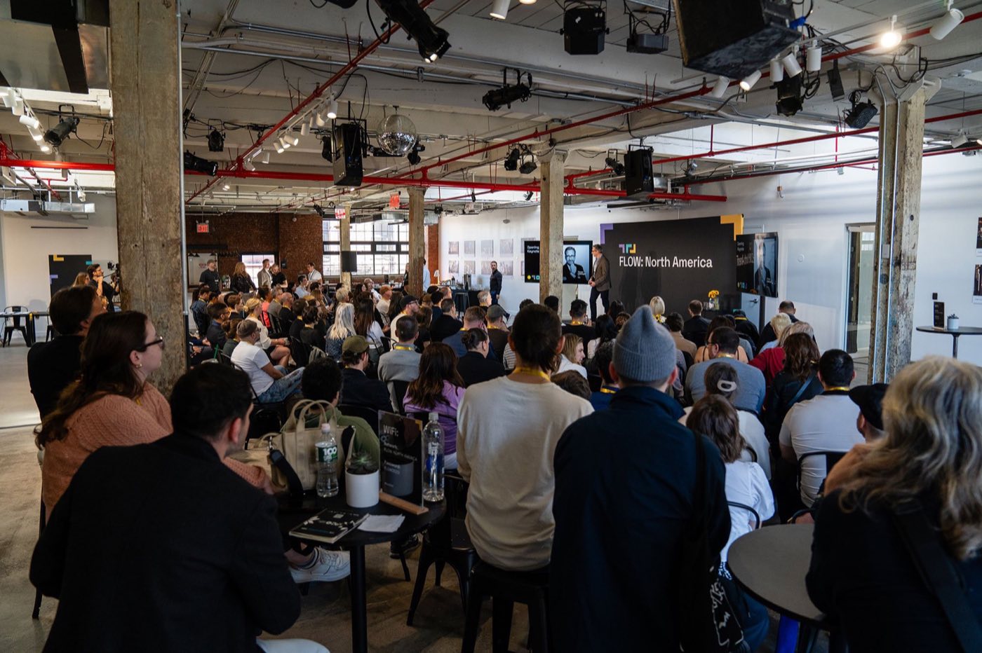 Seated conference audience facing a stage screen in the main industrial loft