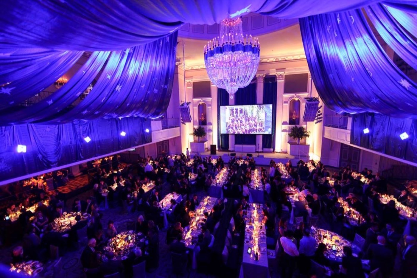 Ballroom draped in blue fabric with a crystal chandelier over candlelit dinner tables
