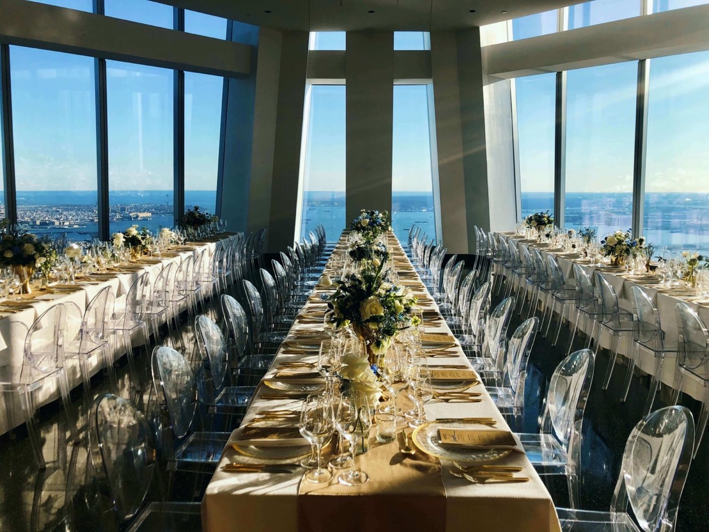 Long banquet tables set for dinner beside floor-to-ceiling windows high above the harbor