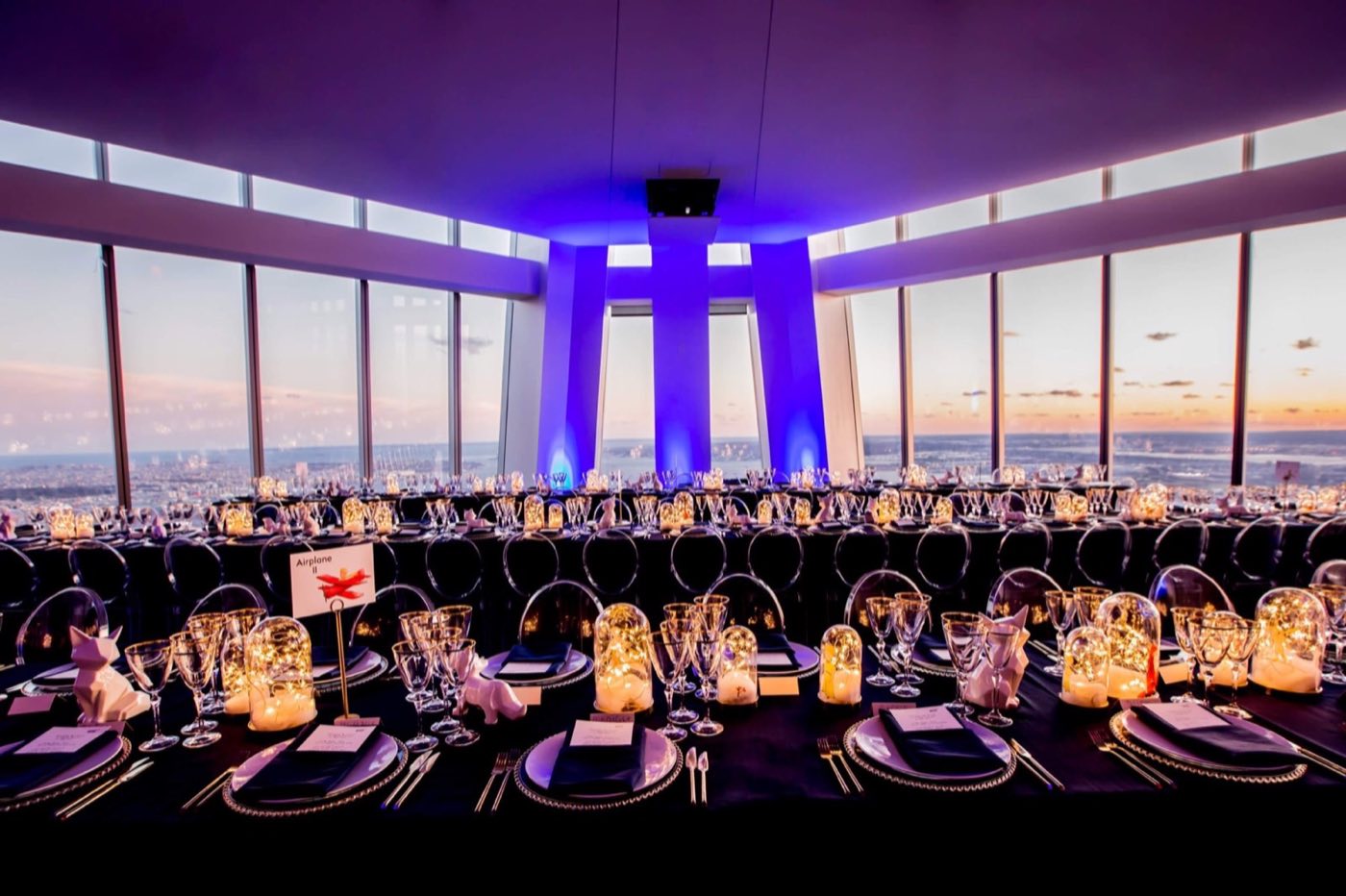 Event floor set for a seated dinner at dusk with purple uplighting and skyline windows