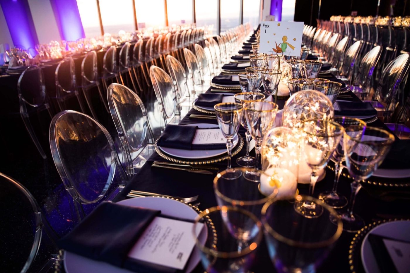 Candlelit dinner tables with clear chairs and glassware under purple lighting