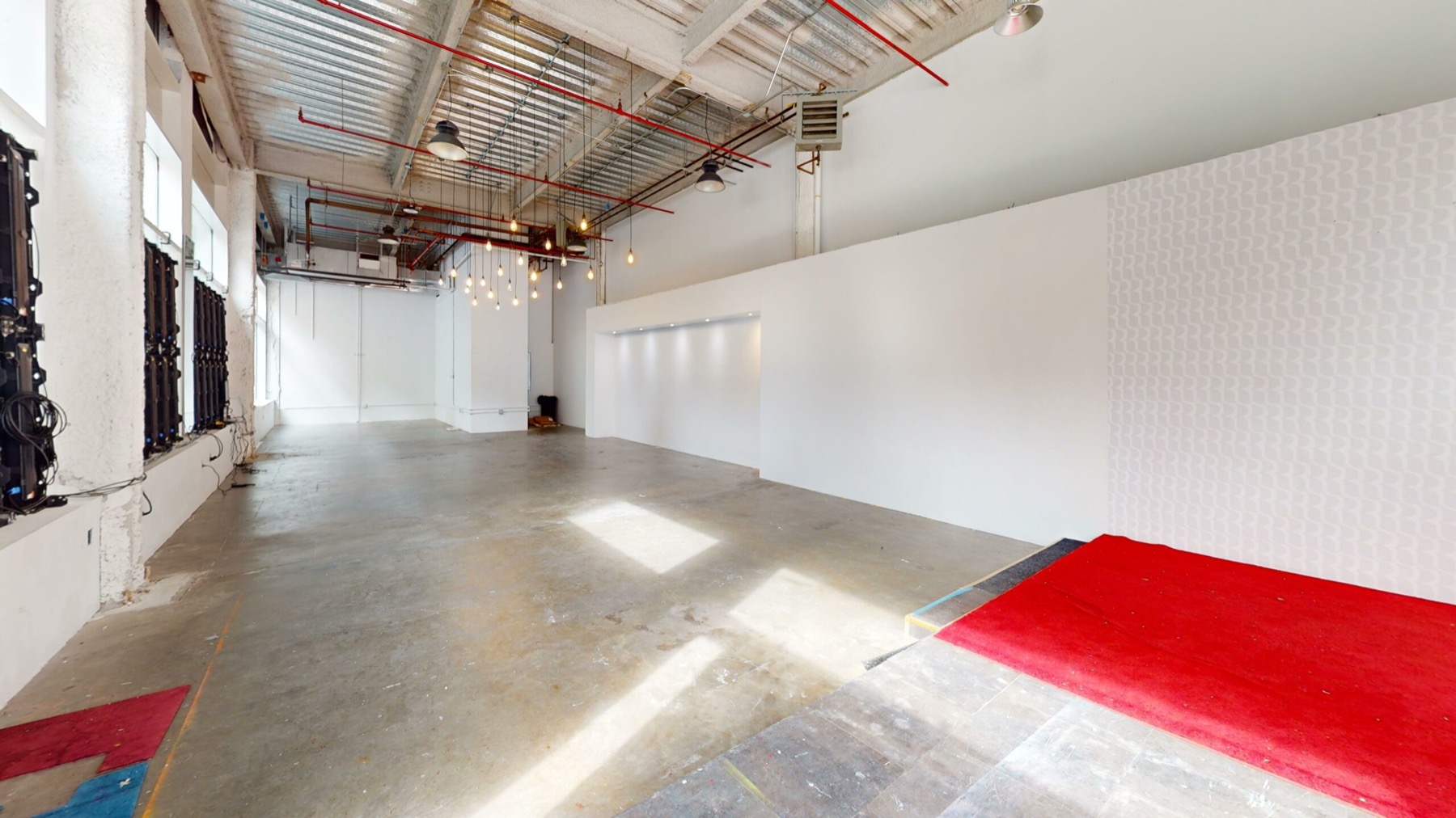Long raw retail room with exposed ceiling pipes, polished concrete and red carpet platform