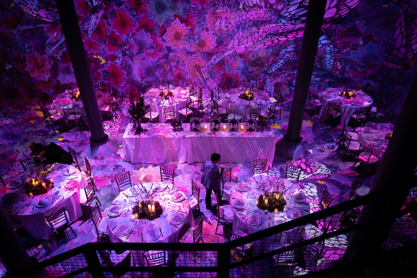 Seated dinner at round tables seen from a balcony, with flower projections on walls and floor