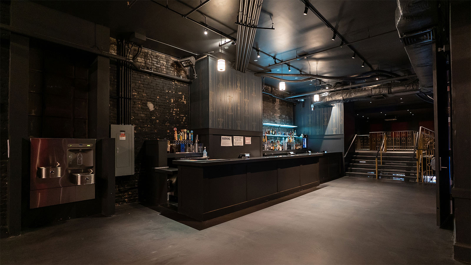 Dark basement bar with exposed brick, industrial ceiling and stairs to the upper level