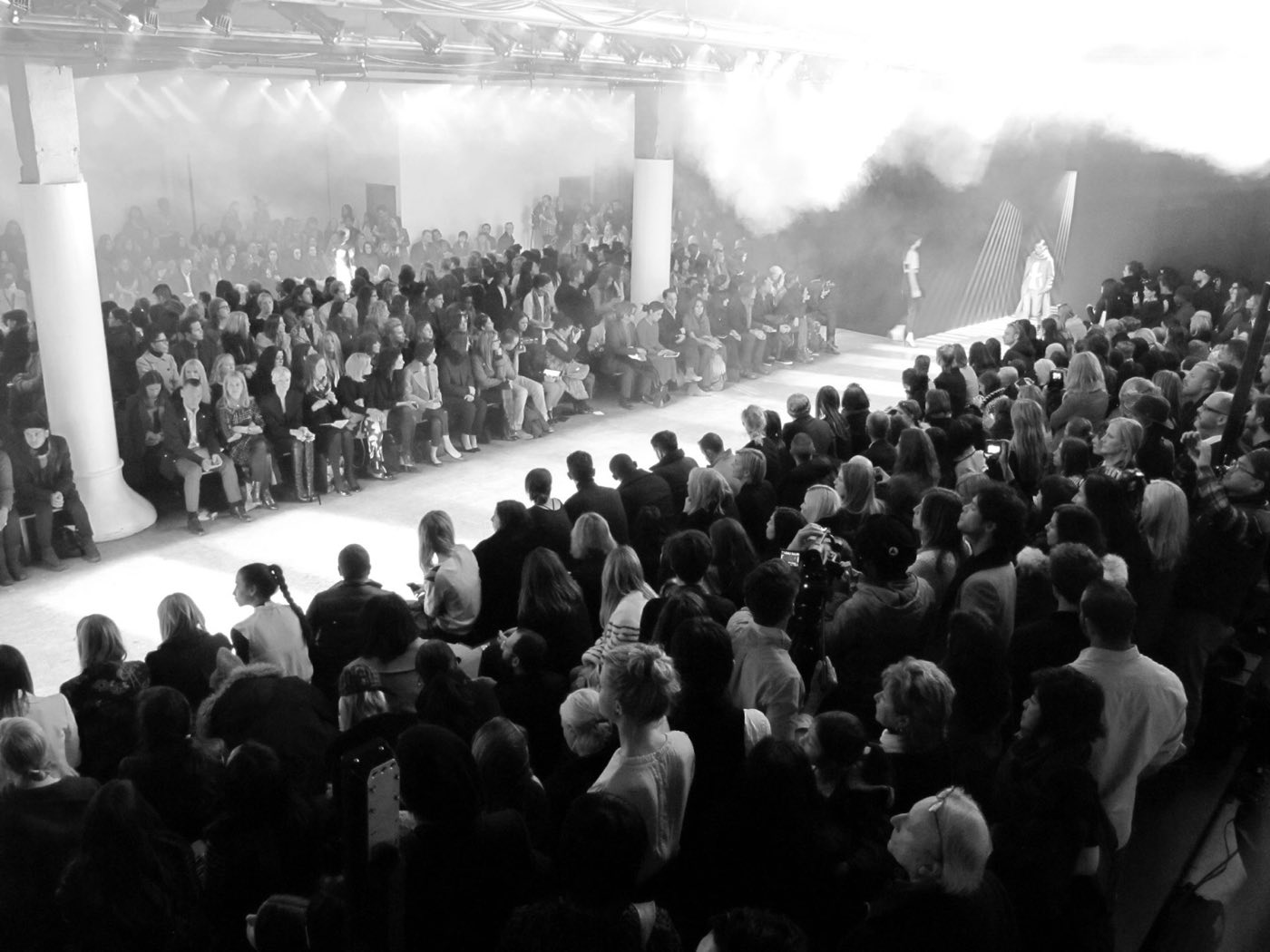 Runway show in a white brick hall with packed seating and haze