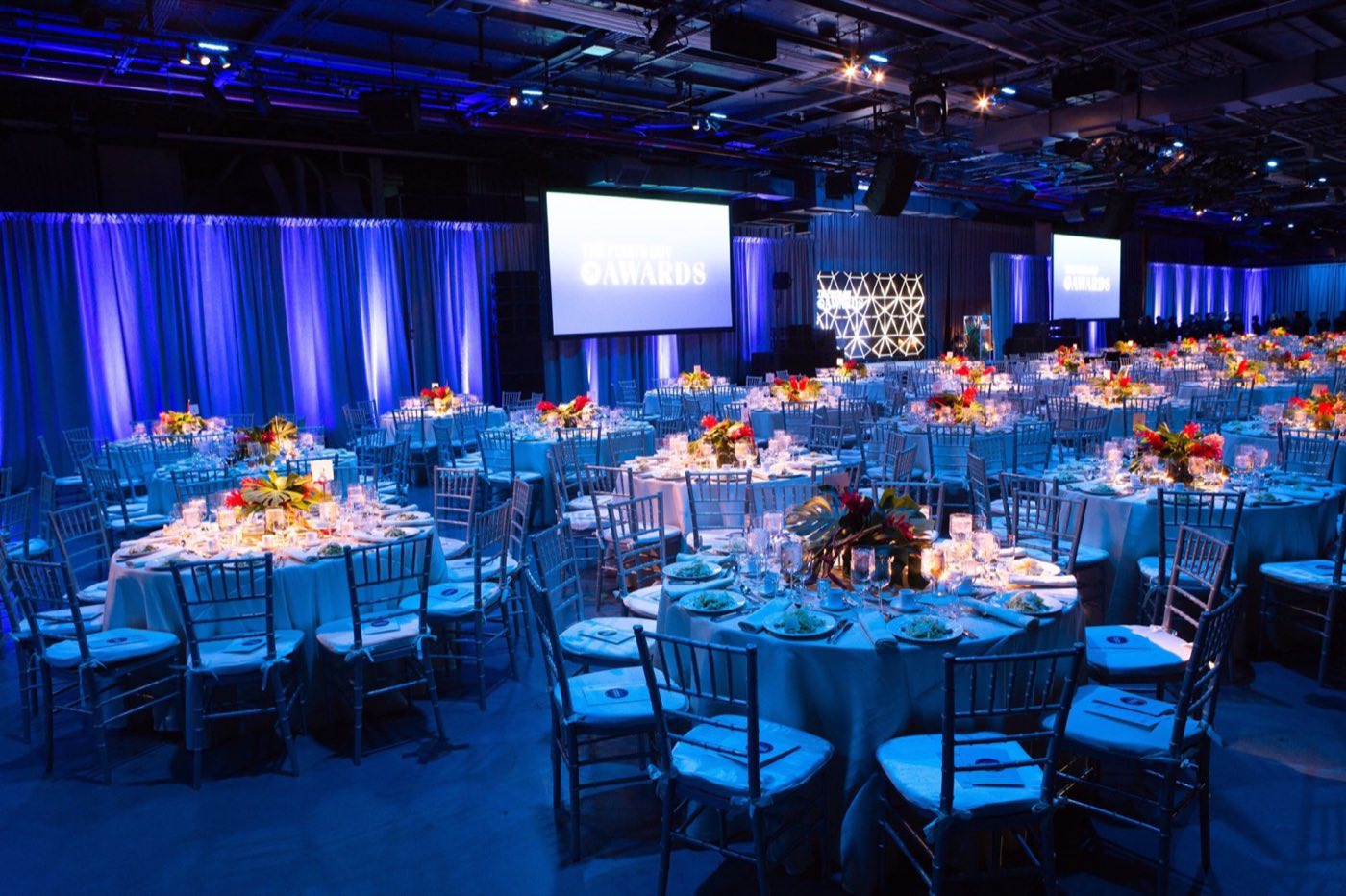 Hangar deck set for a seated gala with round tables, blue drapery and projection screens