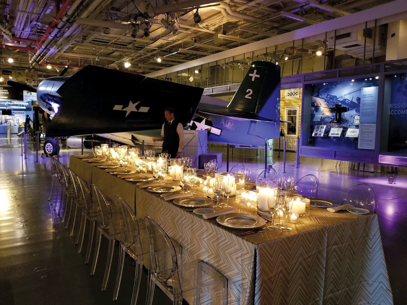 Long candlelit dinner table set beside vintage aircraft on the hangar deck