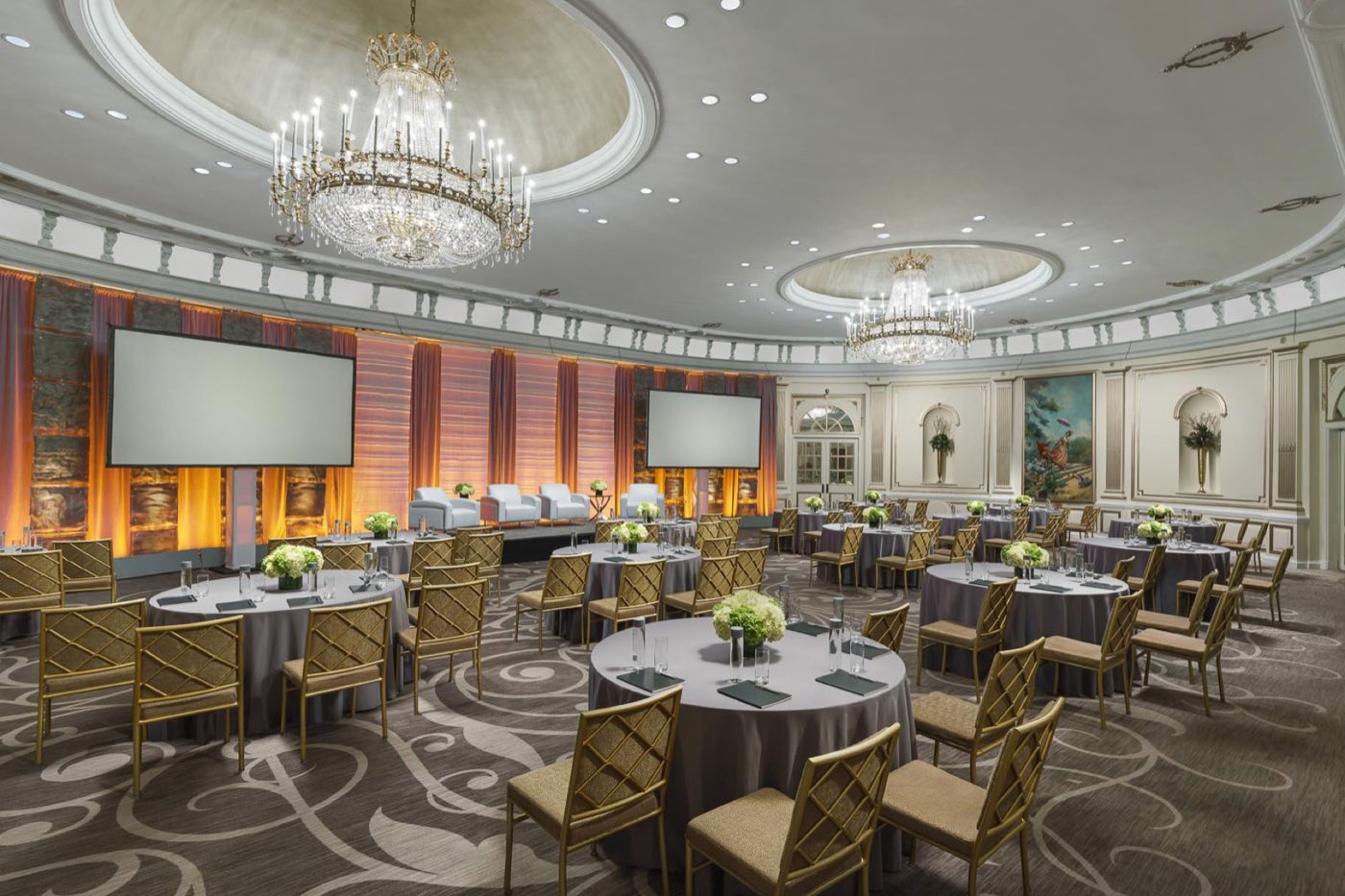 Oval Villard Ballroom set with round tables, two crystal chandeliers and a stage