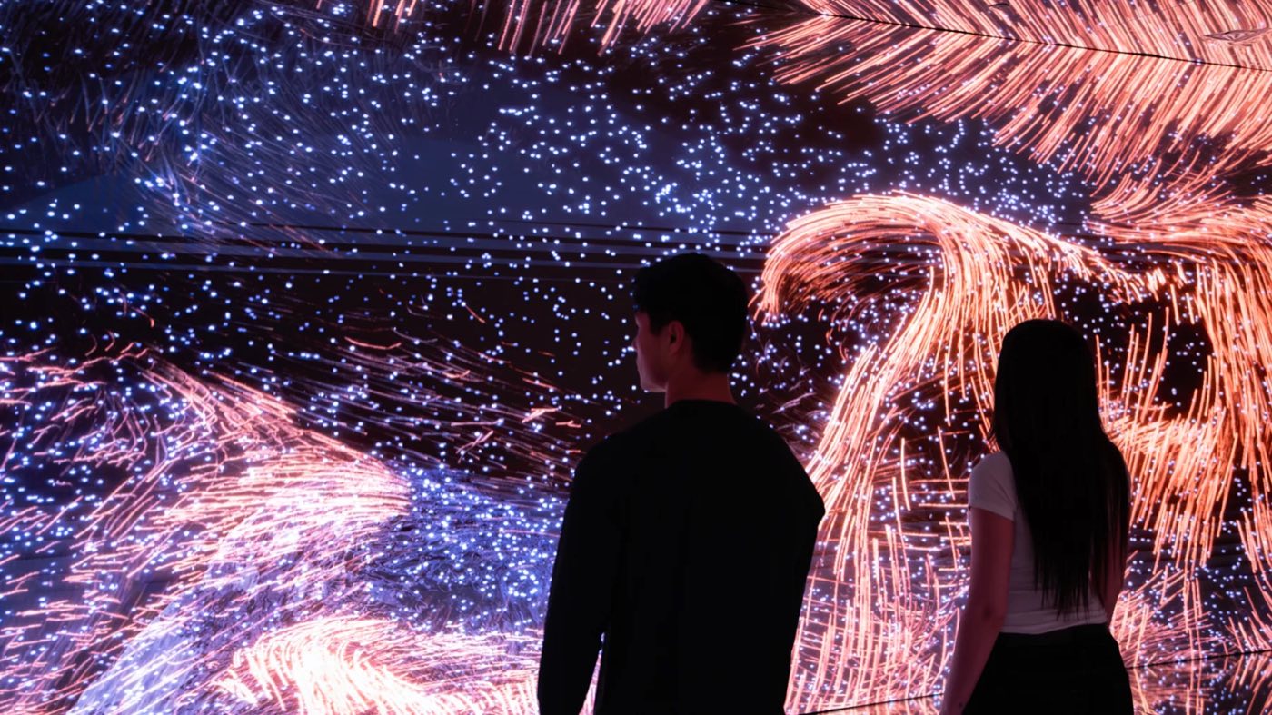 Two visitors in silhouette facing a wall of orange and blue light trails