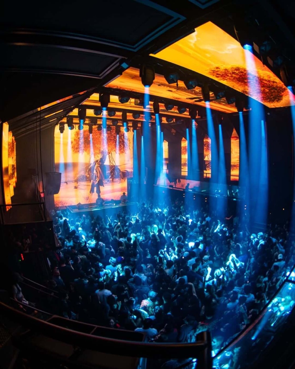 Packed main room during a DJ set, seen from the balcony with blue beam lighting