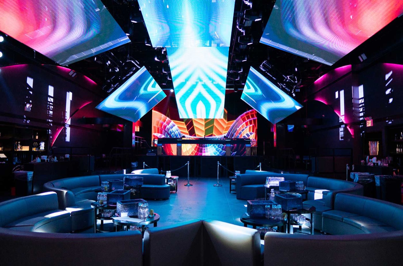 Nightclub floor with curved booths under tilted LED ceiling panels and a lit DJ stage