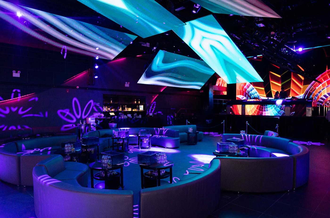 Rounded leather booths on a dark floor beneath angled video panels and a back bar