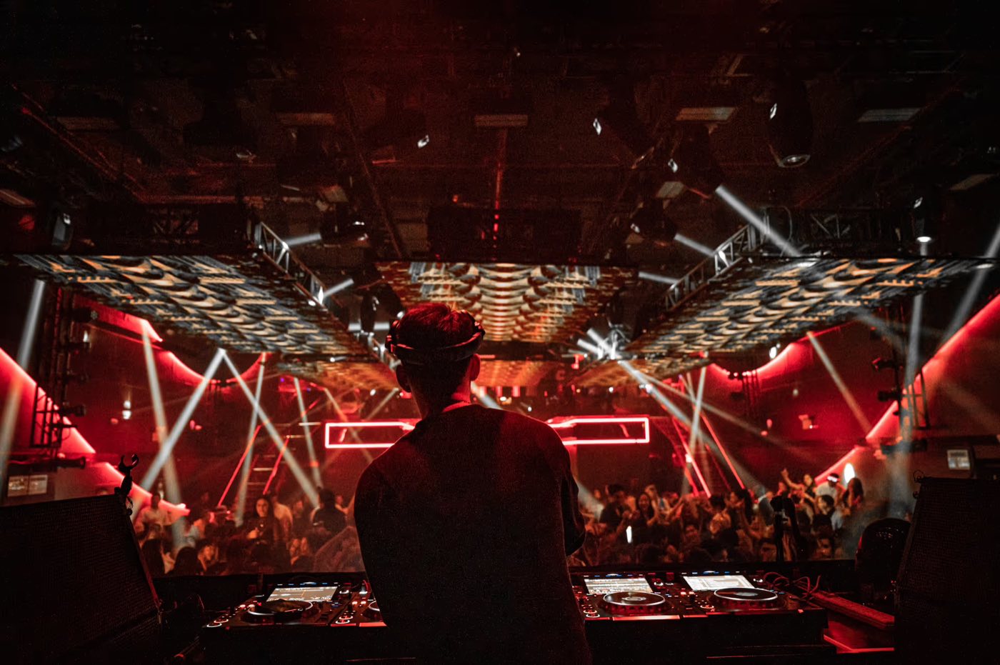 View from behind the DJ booth over a crowd under moving light beams
