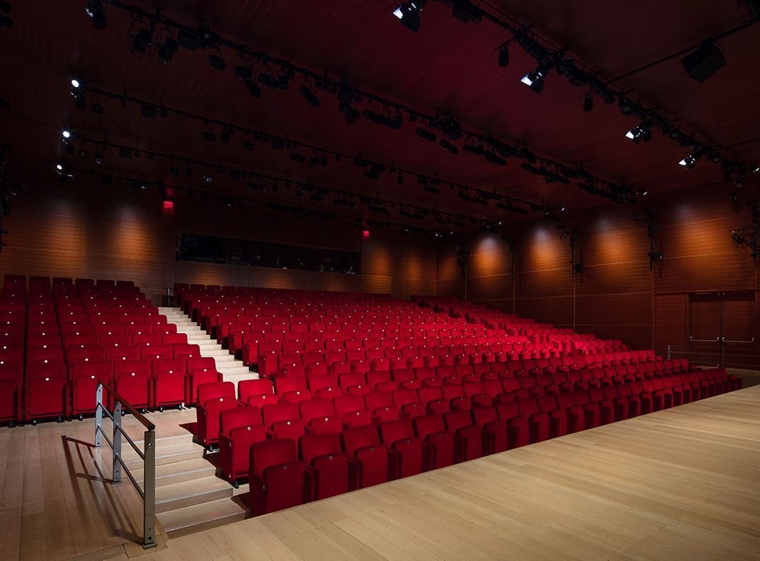 Raked auditorium of red upholstered seats seen from the stage under a lighting grid