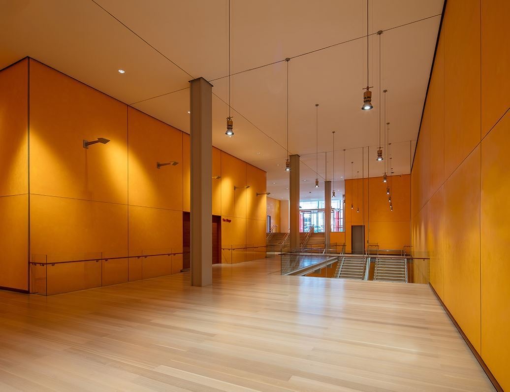 Long open foyer with orange walls, pendant lights and a stairwell down to the stage