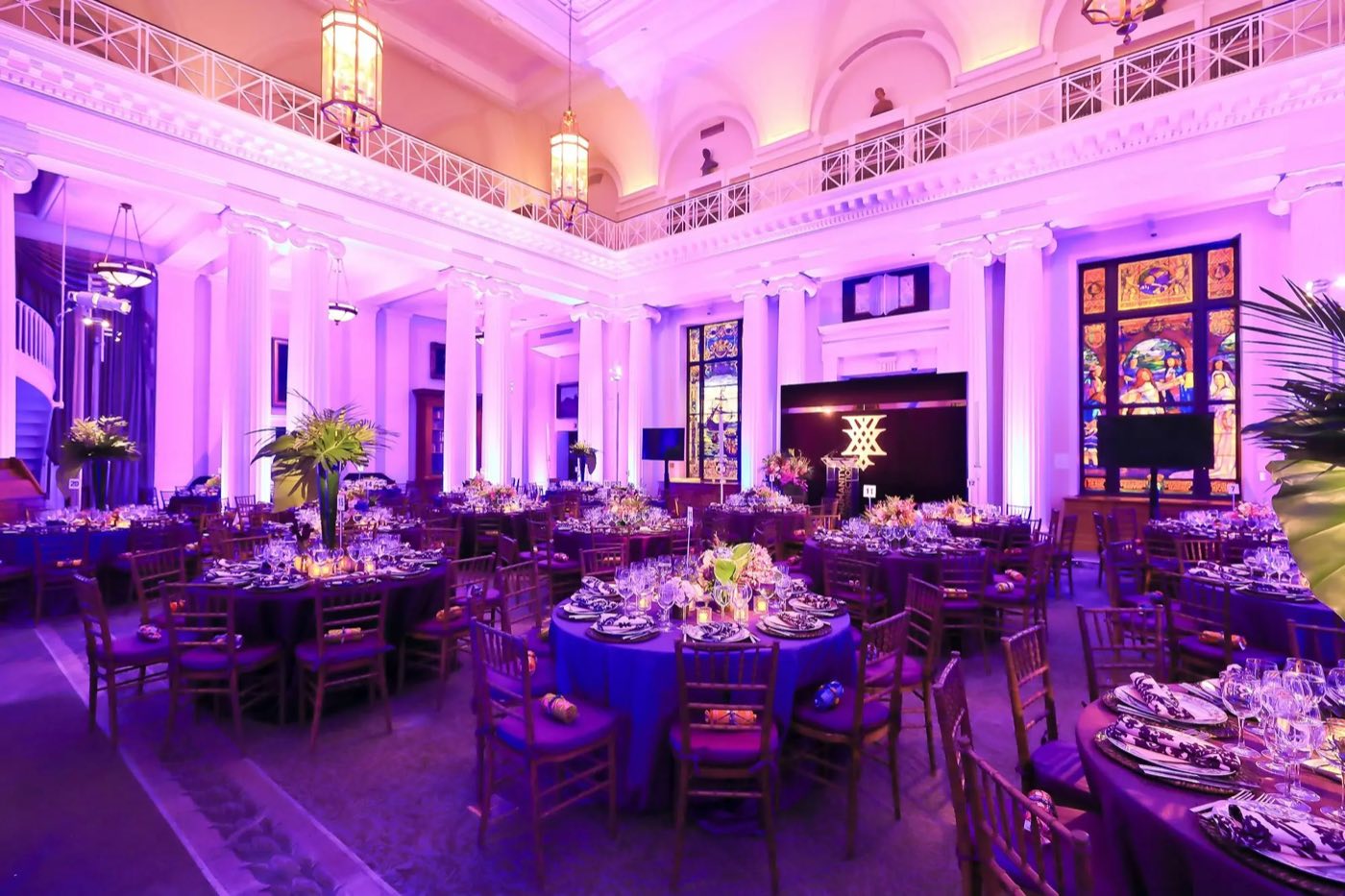 Library set with round dinner tables under violet uplighting and stained-glass windows