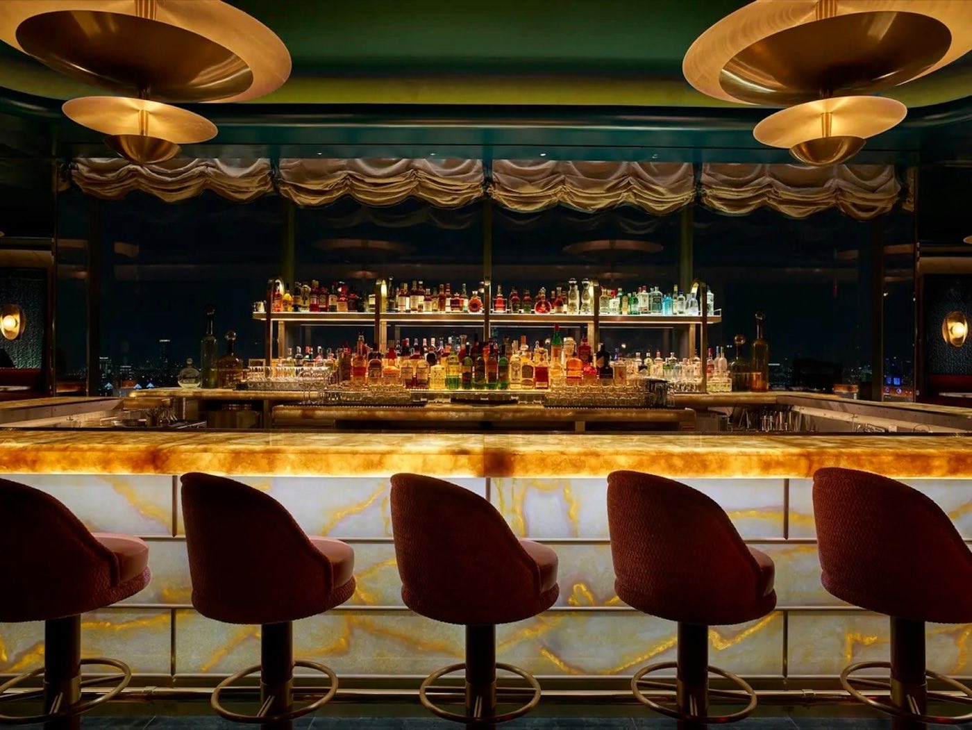 Backlit onyx bar with velvet stools and a mirrored back bar under brass pendants