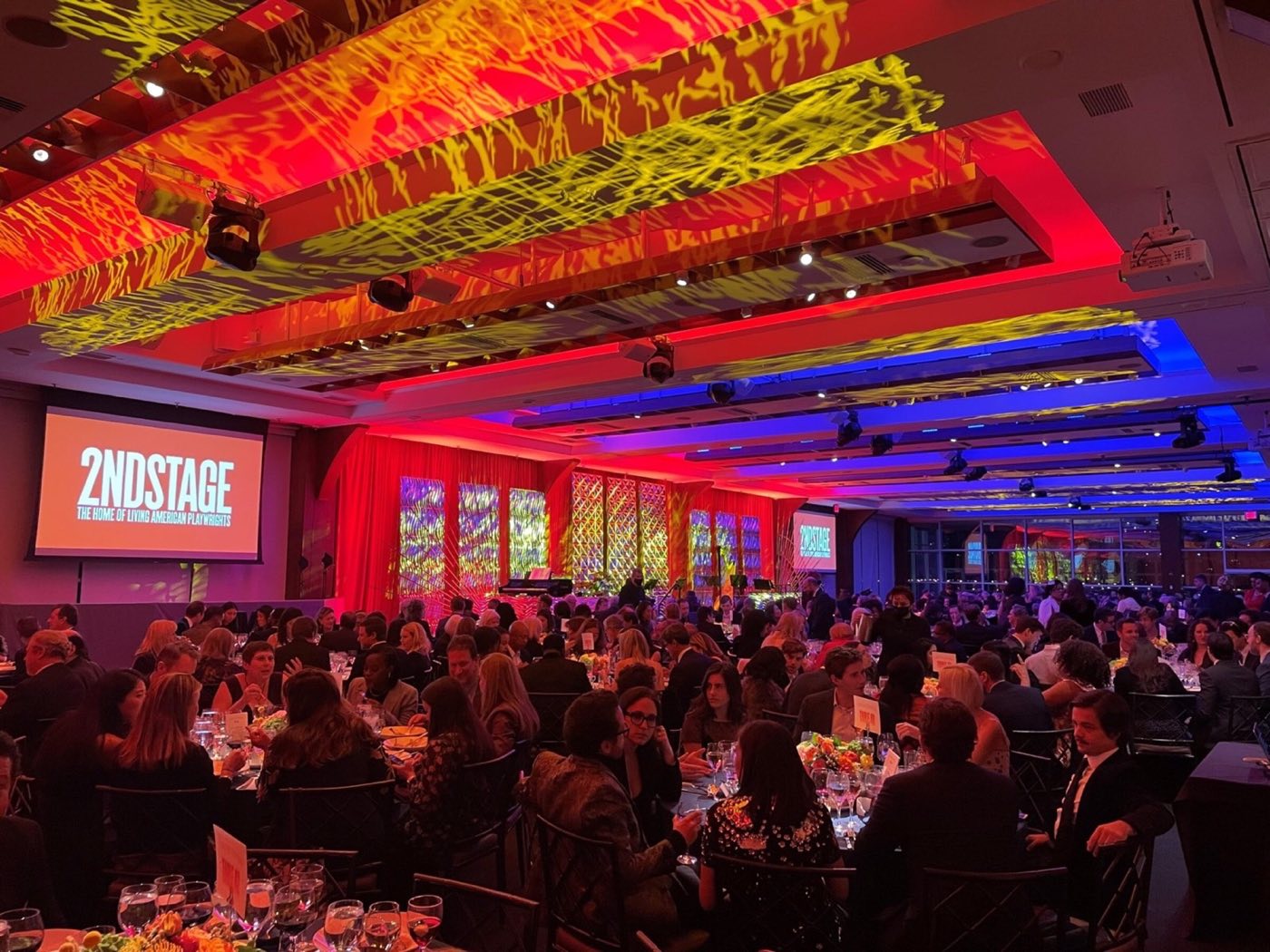 Main ballroom at a seated gala with colored ceiling lighting and a stage screen