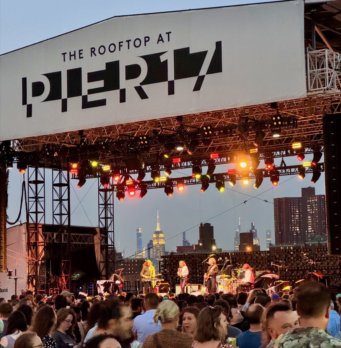 Stage under the rooftop canopy with a crowd facing it at dusk