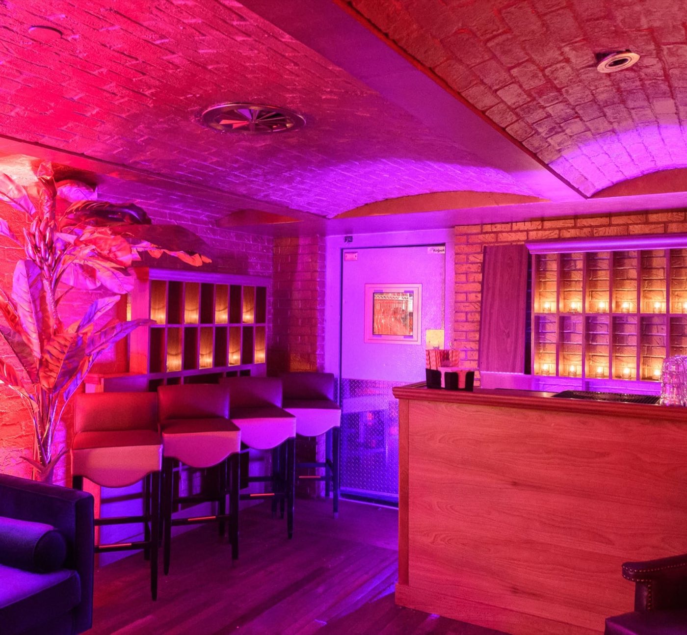 Speakeasy bar with a brick vaulted ceiling, bar stools and a backlit shelf wall