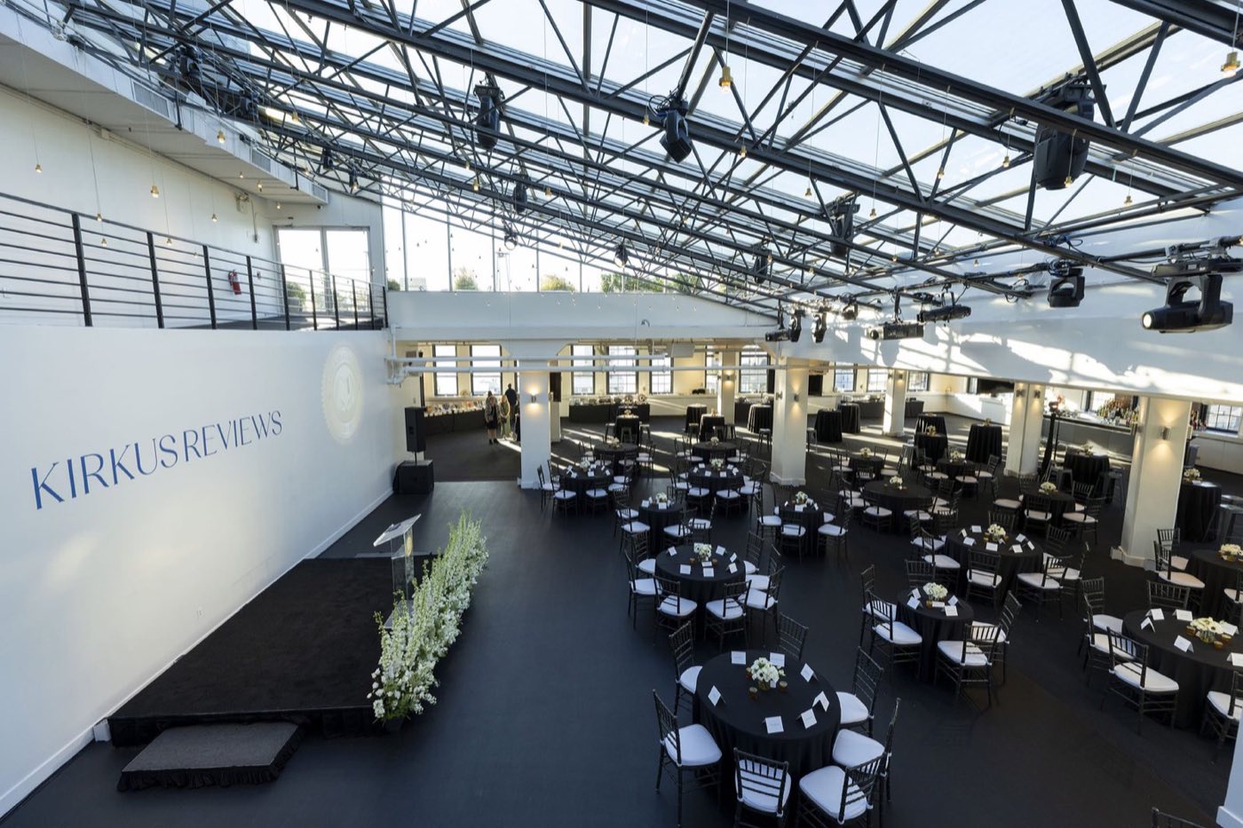 Glass-roofed penthouse hall set with black cocktail tables and a low stage