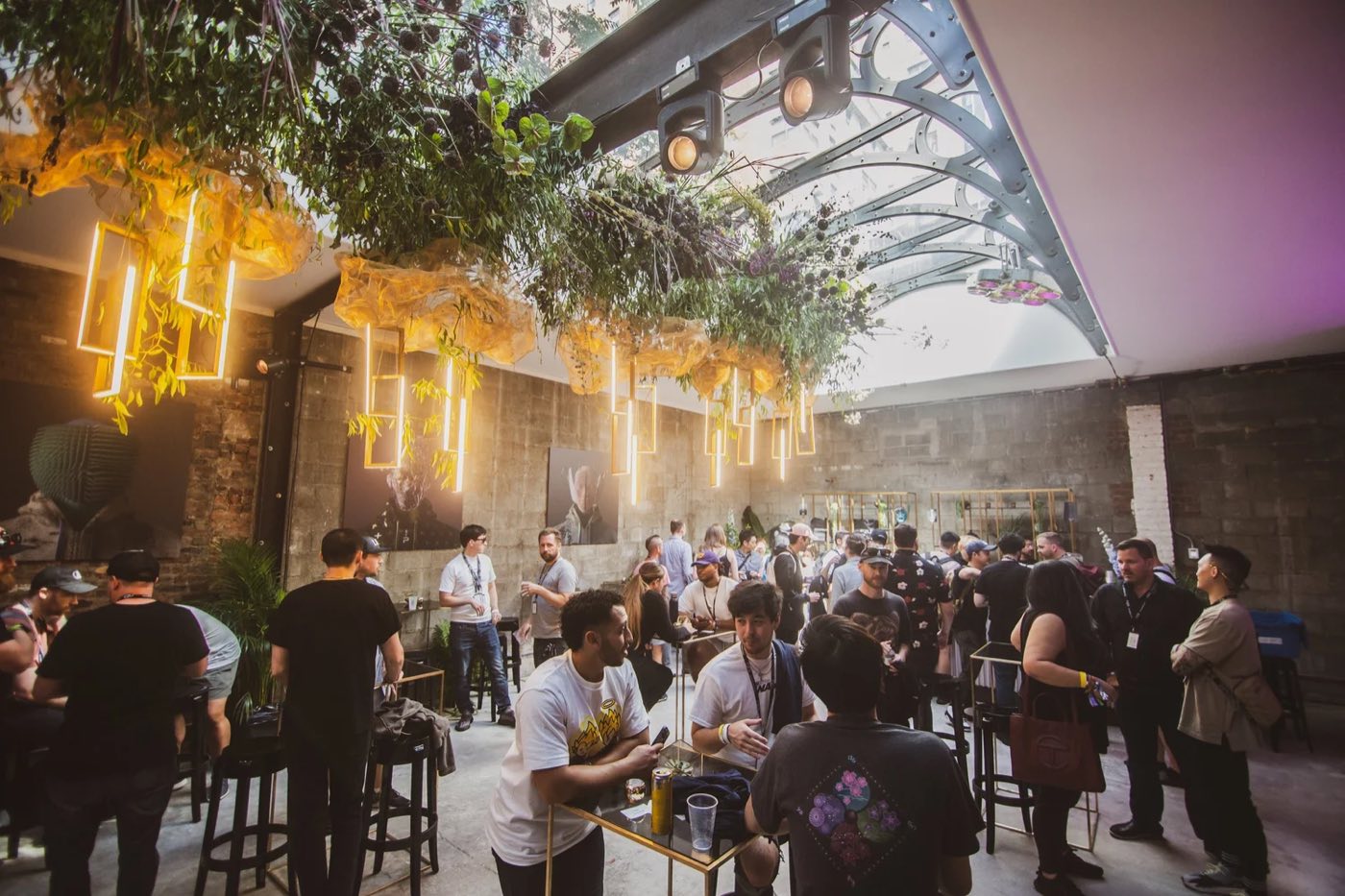 Standing reception under hanging greenery and neon light frames beneath the skylight