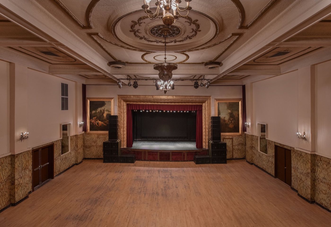 Empty ballroom-style music hall with ornate ceiling, chandeliers and proscenium stage