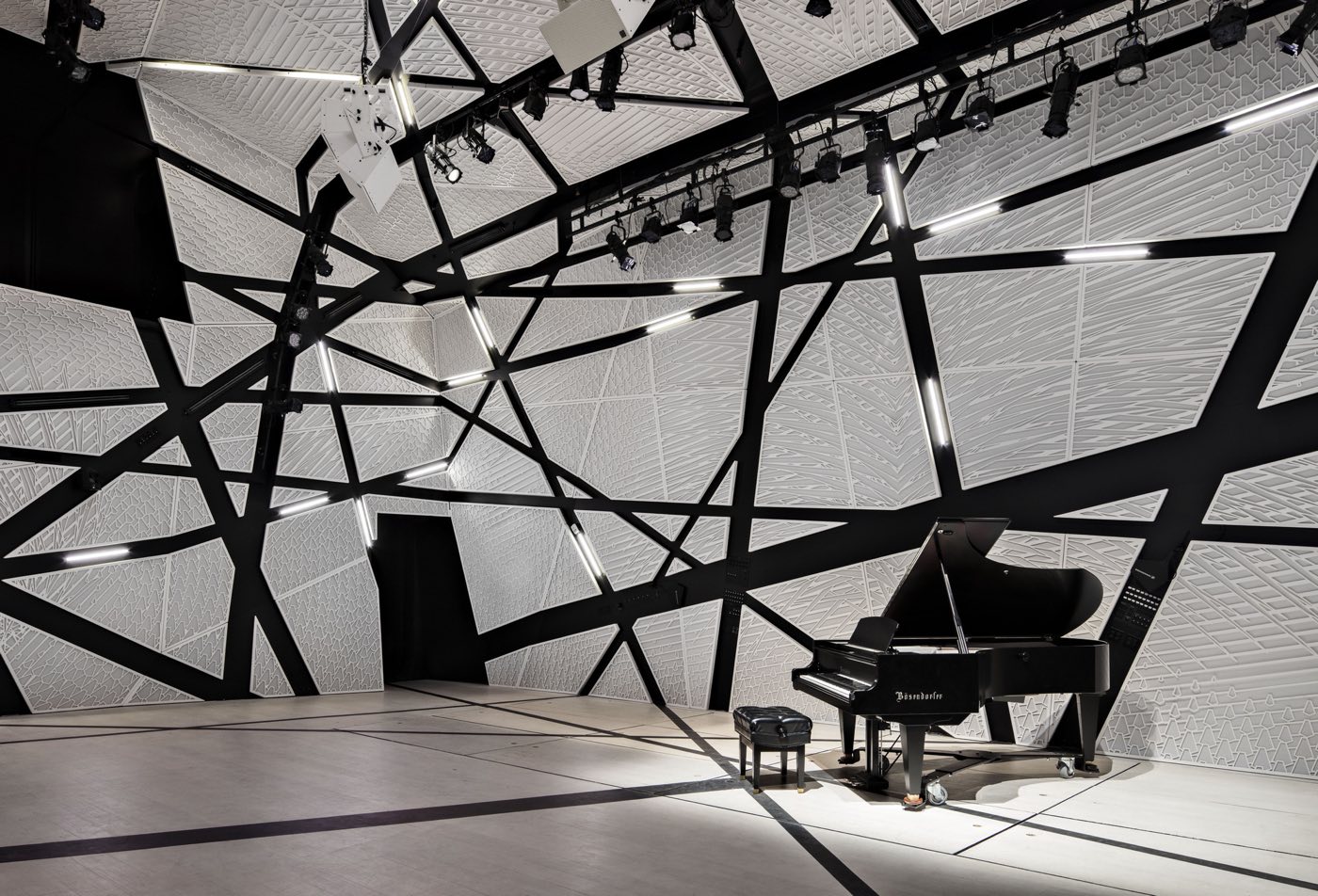 White faceted concert hall with black crisscrossing beams and a grand piano on the floor