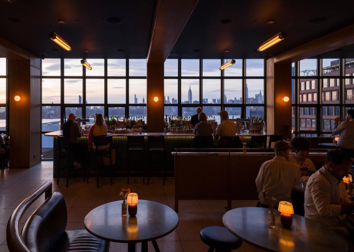 Rooftop bar with cafe tables and a wall of windows facing the Manhattan skyline